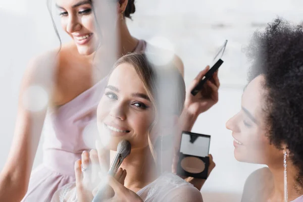 Sposa felice guardando la fotocamera mentre le donne interrazziale applicare il trucco, in primo piano sfocato — Foto stock