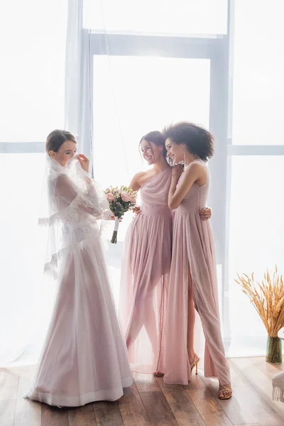 Fröhliche Braut bedeckt Gesicht mit Schleier in der Nähe fröhlicher multikultureller Brautjungfern — Stockfoto