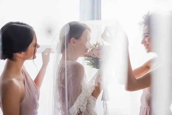 Vue latérale de jolies demoiselles d'honneur interracial près de la mariée souriante voilée au premier plan flou — Photo de stock