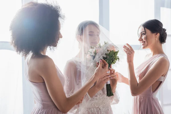 Novia celebración de ramo de boda con los ojos cerrados, mientras que las damas de honor multiculturales que la cubren con velo - foto de stock