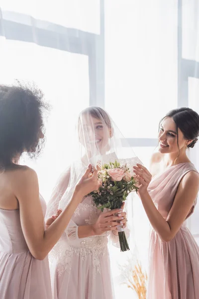 Multicultural bridesmaids touching veil of smiling bride holding wedding bouquet — Stock Photo
