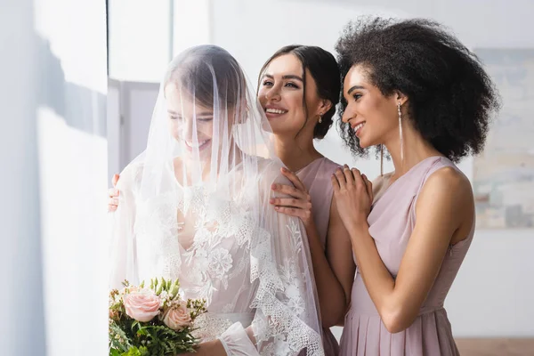 Happy bride holding wedding bouquet near smiling interracial bridesmaids — Stock Photo