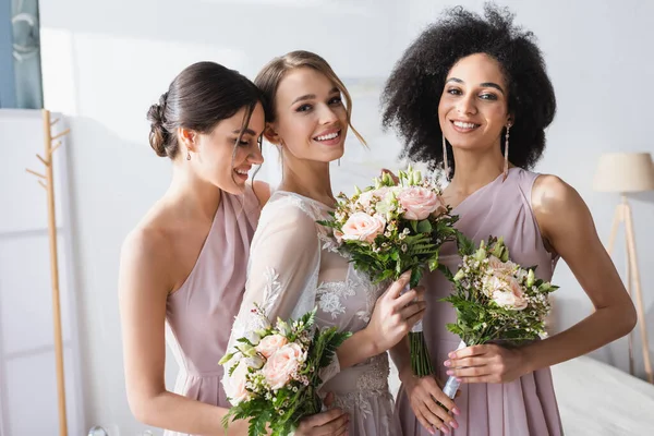 Jolie mariée avec des demoiselles d'honneur interracial tenant des bouquets de mariage dans la chambre — Photo de stock