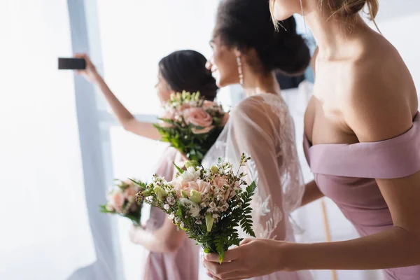 Невеста делает селфи с межрасовыми подружками невесты держа свадебные букеты, размытый фон — стоковое фото