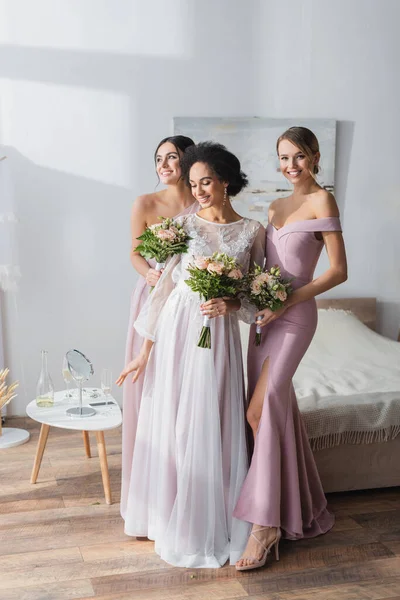 Elegante novia afroamericana con damas de honor felices de pie con ramos de boda en el dormitorio - foto de stock