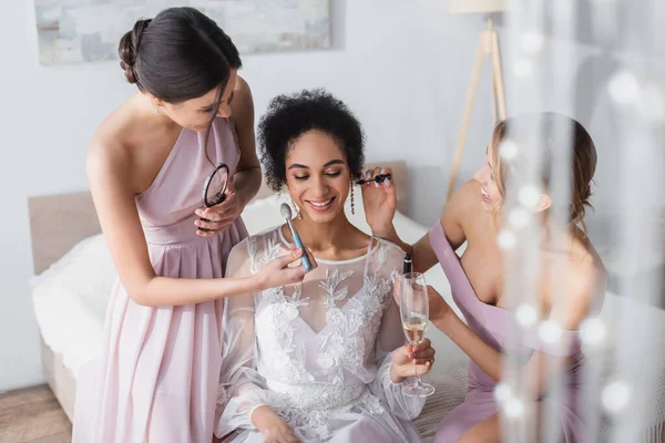 Mujeres jóvenes haciendo maquillaje a la novia afroamericana feliz en el dormitorio - foto de stock