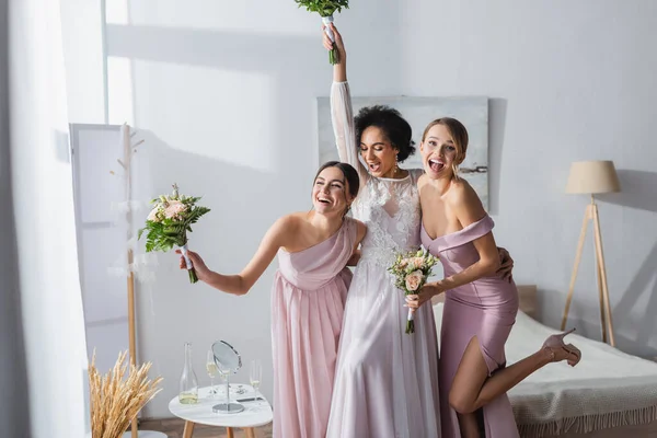 Excitée mariée afro-américaine avec demoiselles d'honneur tenant des bouquets de mariage tout en s'amusant dans la chambre — Photo de stock