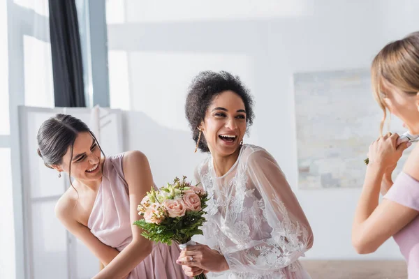 Aufgeregte afrikanisch-amerikanische Braut hält Hochzeitsstrauß und lacht in der Nähe von Brautjungfern — Stockfoto