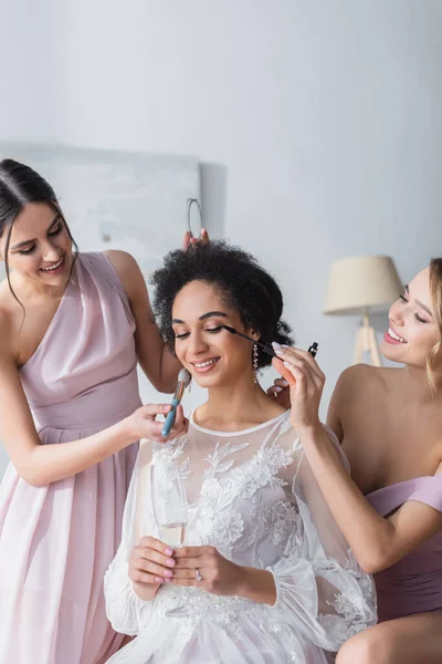 Damas de honra sorridentes aplicando rímel e pó facial na noiva afro-americana — Fotografia de Stock