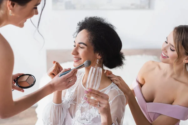 African american bride holding champagne while bridesmaid applying highlighter, blurred foreground — Stock Photo