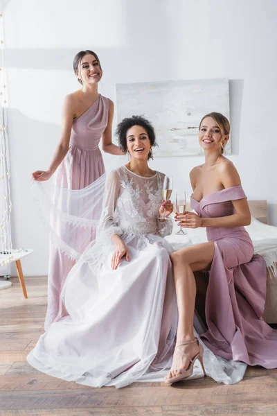 Feliz afroamericana novia con damas de honor sonriendo a la cámara en el dormitorio - foto de stock