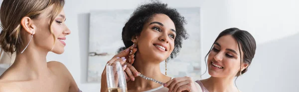 Joven dama de honor sosteniendo collar cerca de la novia afroamericana feliz, bandera - foto de stock