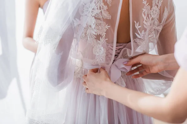 Partial view of bridesmaid tying ribbon on lace dress of bride — Stock Photo