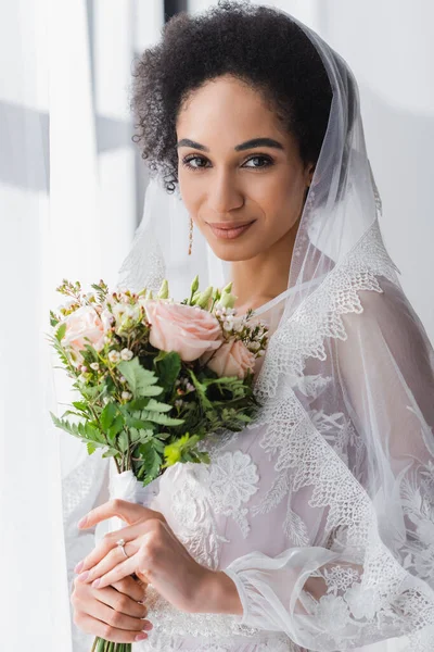 Jolie mariée afro-américaine tenant bouquet de mariage tout en regardant caméra — Photo de stock