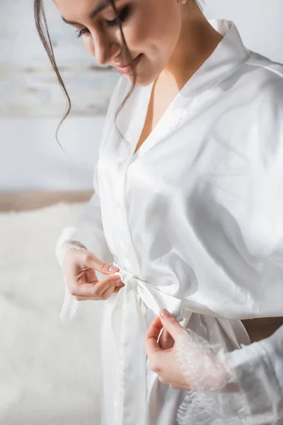 Young bride tying white satin robe in bedroom — Stock Photo