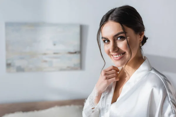 Happy, brunette bride in white robe looking at camera in bedroom — Stock Photo