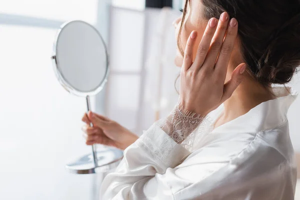 Young bride holding mirror and fixing hair in bedroom — Stock Photo