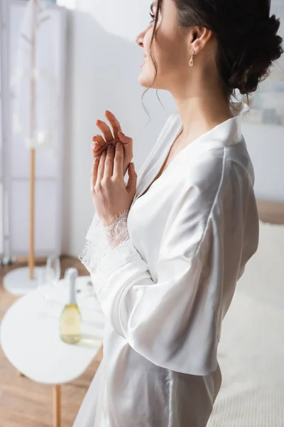 Smiling and dreamy bride in white robe looking away in bedroom — Stock Photo