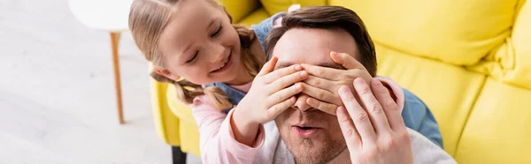 Niño feliz cubriendo los ojos del padre sorprendido mientras se divierte en casa, pancarta - foto de stock