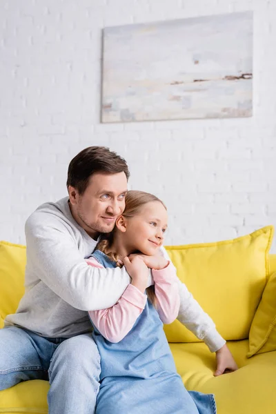 Lächelnder Vater schaut weg, während er seine Tochter zu Hause auf dem Sofa umarmt — Stockfoto