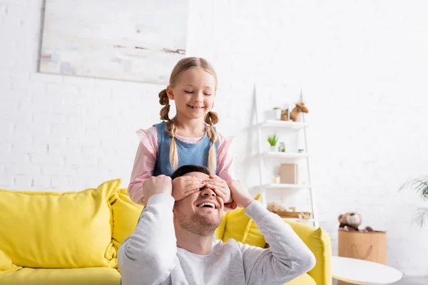 Улыбающаяся девушка закрывает глаза смеющегося отца, веселясь дома — стоковое фото