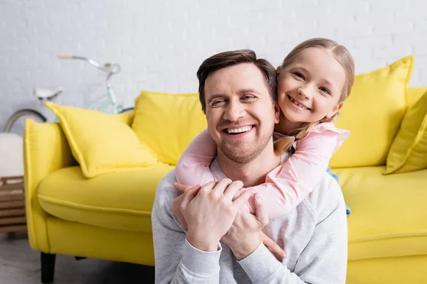 Adult man laughing while cheerful daughter hugging him at home — Stock Photo