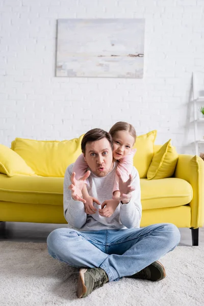 Hombre sentado en el suelo y mostrando gesto aterrador cerca de hija alegre - foto de stock