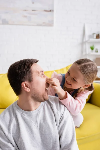 Chica juguetona taponamiento nariz de padre mientras se divierten en la sala de estar - foto de stock