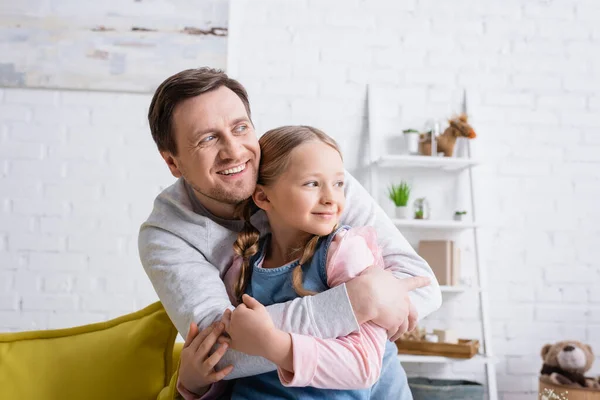 Menina alegre com o pai olhando para longe enquanto abraçando em casa — Fotografia de Stock