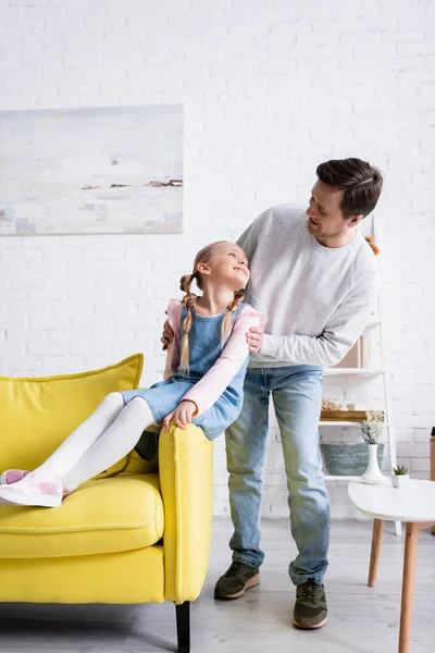 Hombre feliz tocando hombros de hija alegre sentada en el sofá en la sala de estar - foto de stock