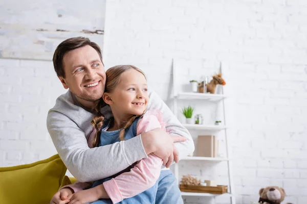Sonriente hombre mirando hacia otro lado mientras abraza a su hija en casa - foto de stock