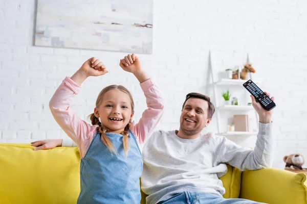 Cheerful girl showing triumph gesture while watching tv near dad at home — Stock Photo