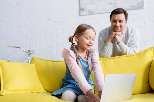 Joyful man looking at daughter using laptop on sofa — Stock Photo