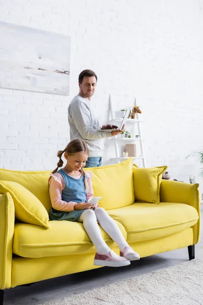 Lächelndes Mädchen sitzt auf Sofa mit Smartphone neben Vater mit Laptop — Stockfoto