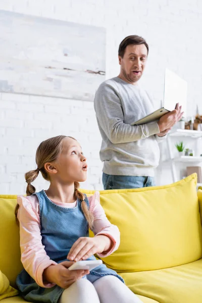 Amazed man with laptop looking at surprised daughter using smartphone on sofa — Stock Photo