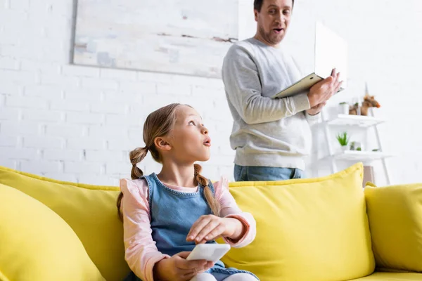 Surprised man with laptop looking at daughter covering smartphone with hand — Stock Photo