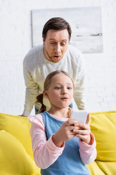Изумленный отец рядом дочь болтает на смартфоне дома — стоковое фото