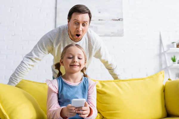 Surprised man looking at smartphone in hands of daughter sitting on sofa — Stock Photo