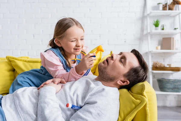 Mädchen hält Spielzeug-Otoskop neben Vater, der mit geschlossenen Augen auf Sofa liegt — Stockfoto