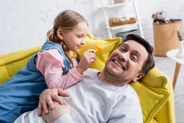 Alegre hombre acostado en sofá cerca de hija sosteniendo otoscopio de juguete — Stock Photo