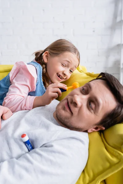 Мужчина лежит на диване рядом с дочерью, осматривая его с помощью отоскопа, играя в доктора, размытый передний план — стоковое фото