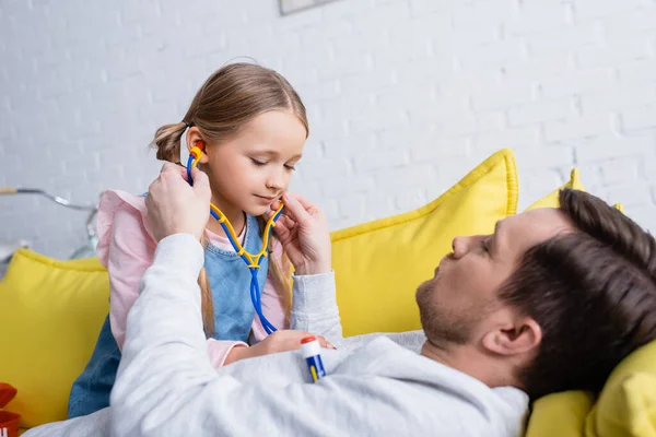 Man lying on sofa while daughter examining him with toy stethoscope — Stock Photo
