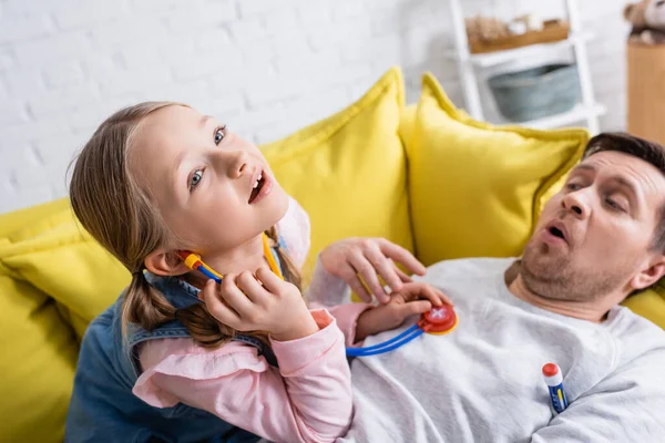 Мужчина лежит на диване и притворяется больным, пока дочь осматривает его с помощью игрушечного стетоскопа — стоковое фото
