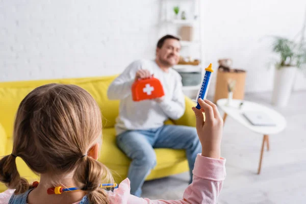 Vista posteriore del bambino con siringa giocattolo vicino spaventato padre tenendo kit di pronto soccorso su sfondo sfocato — Foto stock