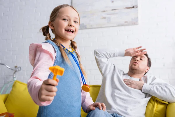 Excitada chica sosteniendo juguete reflejo martillo mientras jugando médico cerca papá fingiendo enfermo sobre fondo borroso - foto de stock