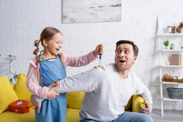 Excitada chica haciendo inyección con jeringa de juguete a padre fingiendo asustado - foto de stock