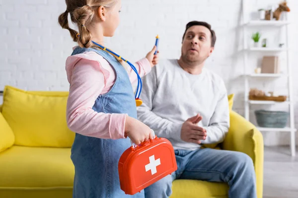 Girl holding toy syringe and first aid kit near father pretending scared on blurred background — Stock Photo