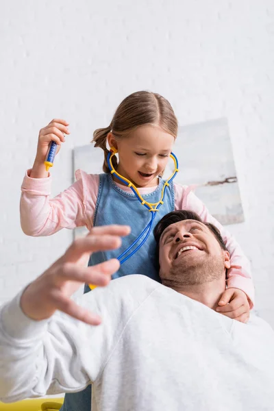 Menina segurando brinquedo seringa perto pai fingindo assustado em primeiro plano borrado — Fotografia de Stock