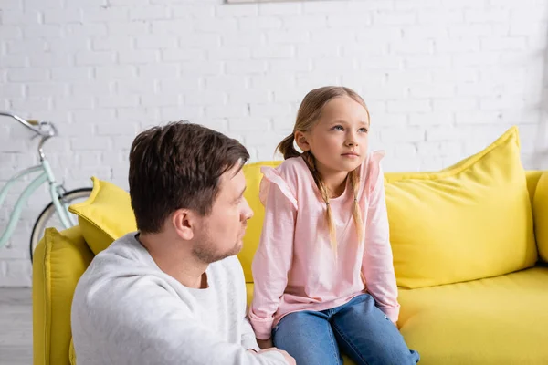 Bouleversée fille regardant loin tout en étant assis sur le canapé près du père — Photo de stock