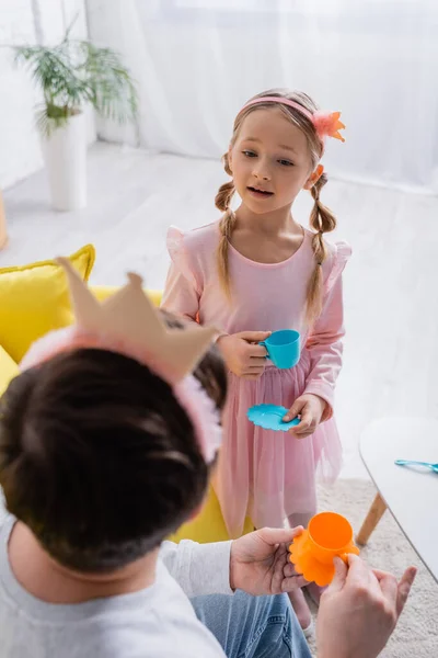 Вид сзади мужчины в игрушечной короне рядом с дочерью, играющей дома, размытый передний план — стоковое фото